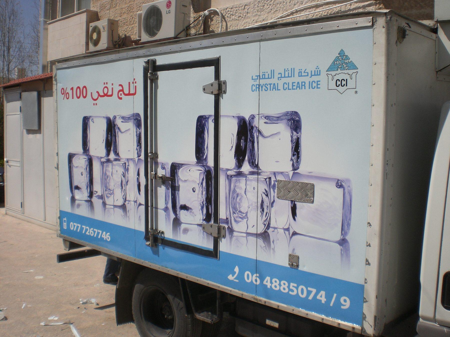 Side of a white box truck with ice-cube artwork, Arabic text and phone numbers.