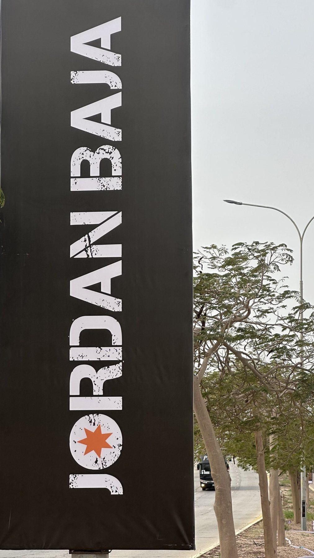 A tall black vertical banner with white lettering stands on a roadside pole.