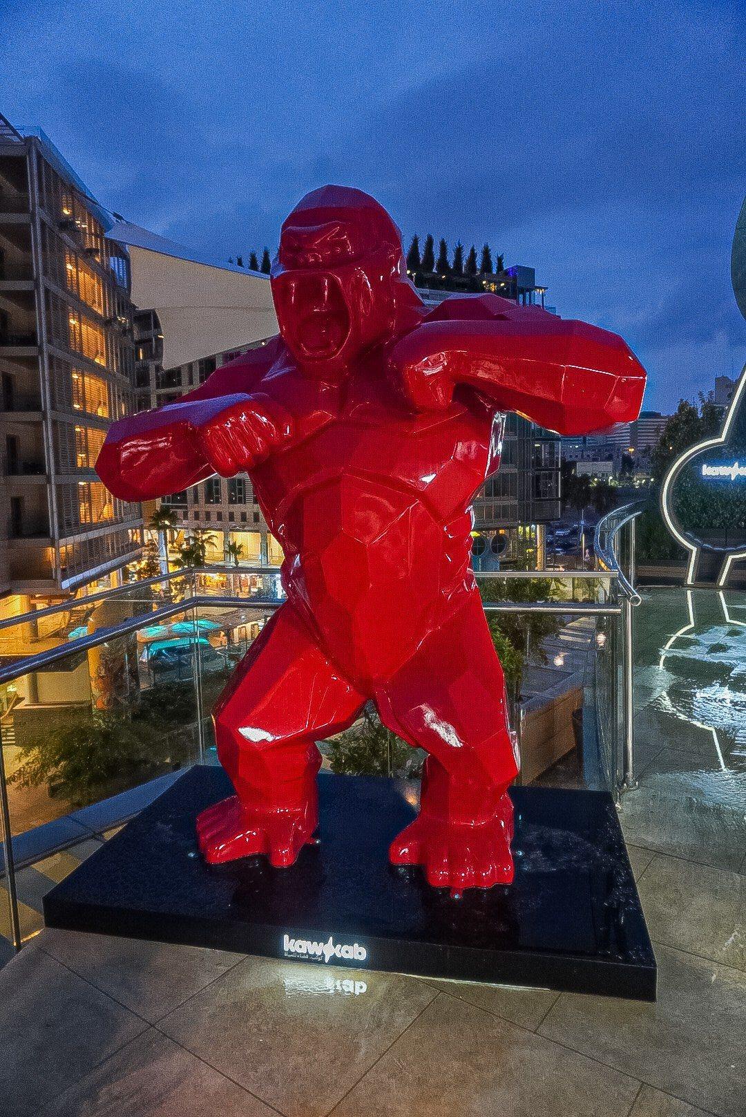 A large glossy red gorilla sculpture on a black plinth outdoors at night.