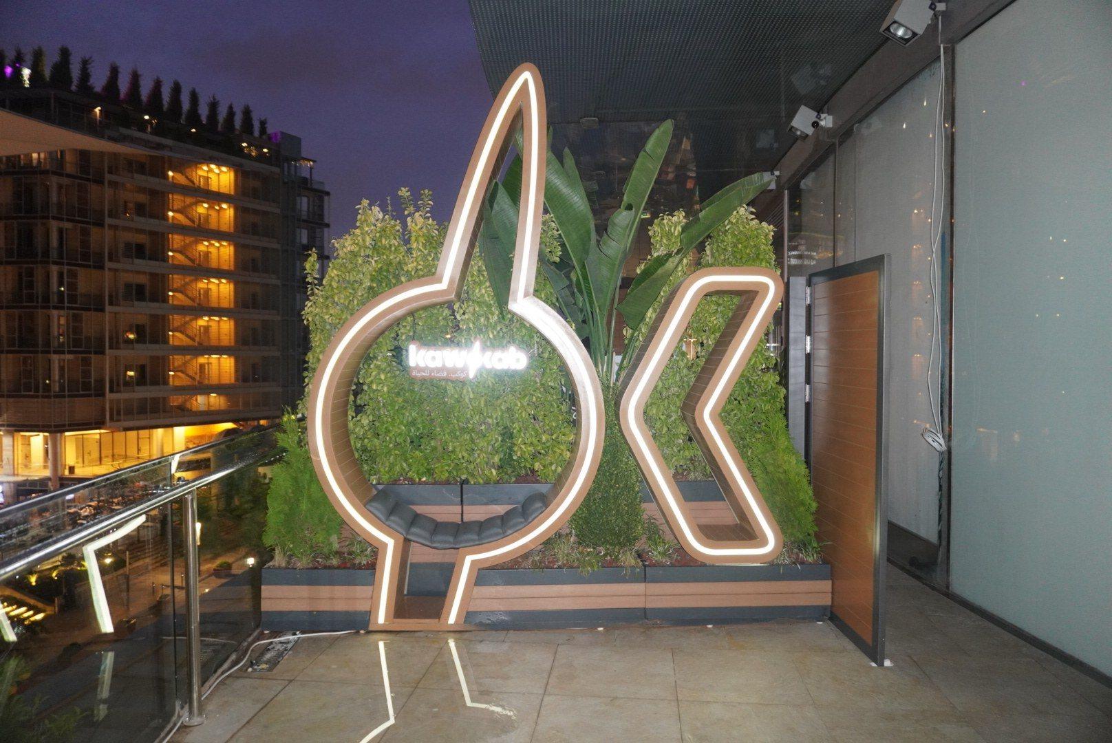 An illuminated neon logo sign set among plants in an atrium at night.
