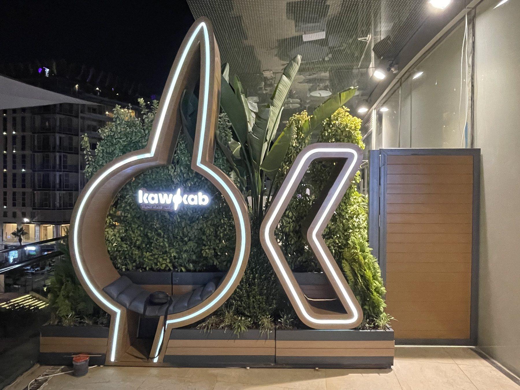 Large illuminated outline logo with a curved shape and letters, mounted above planters at night.