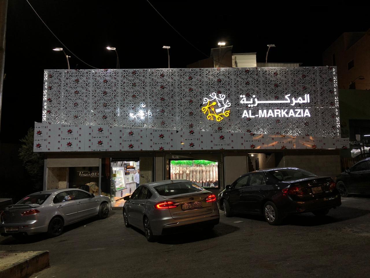 Illuminated lettering and logo on a patterned facade at night, with cars parked in front.
