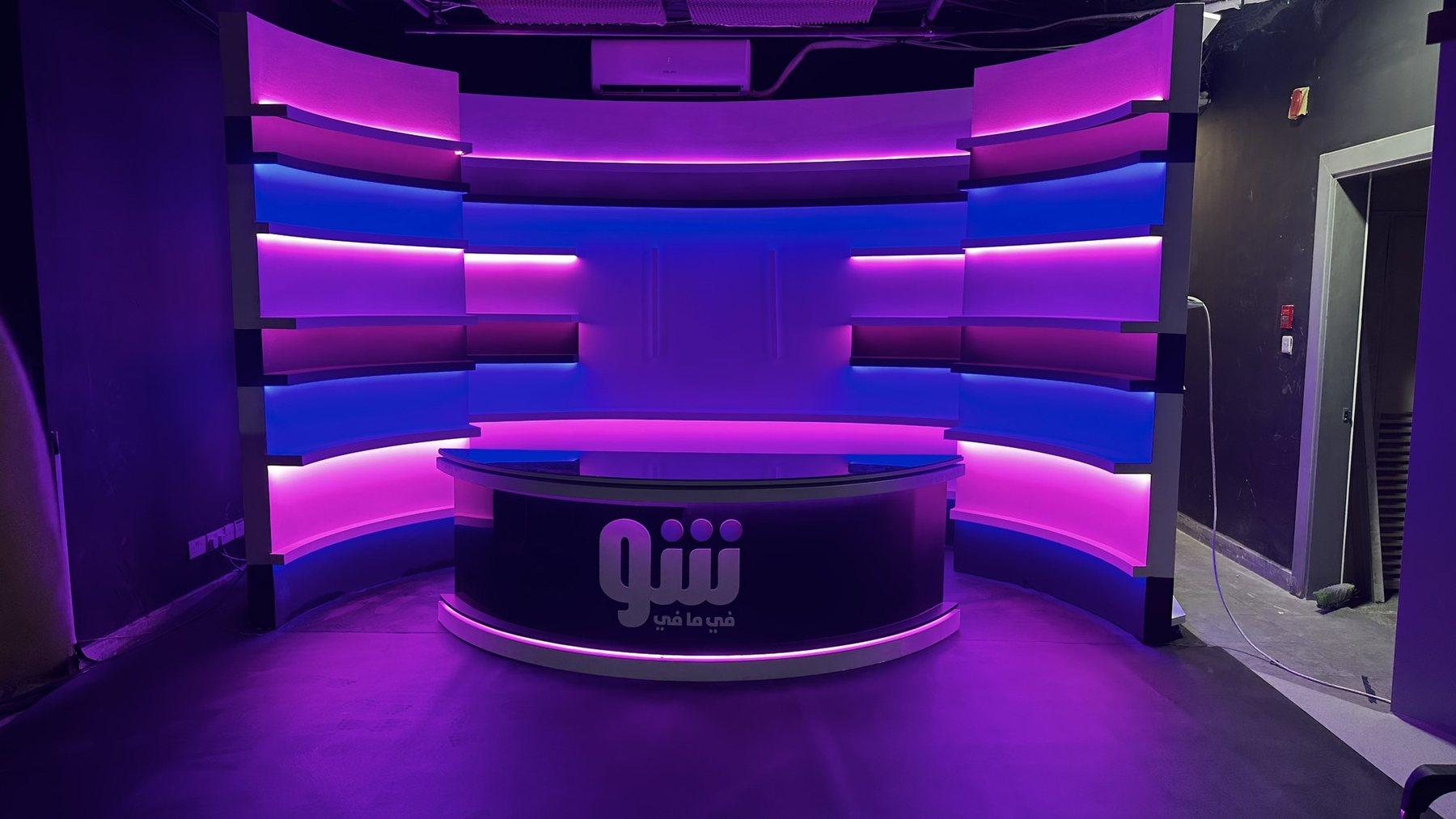 A purple and magenta lit TV studio with a curved presenter desk and wall LED strips.
