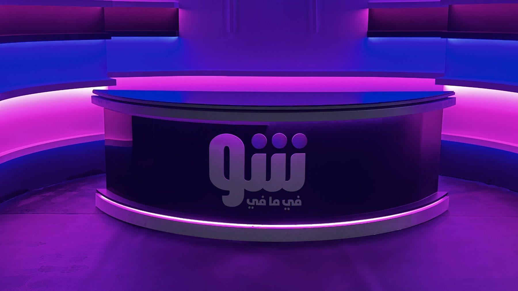 A curved presenter desk with a back-lit logo inside a purple-lit studio.