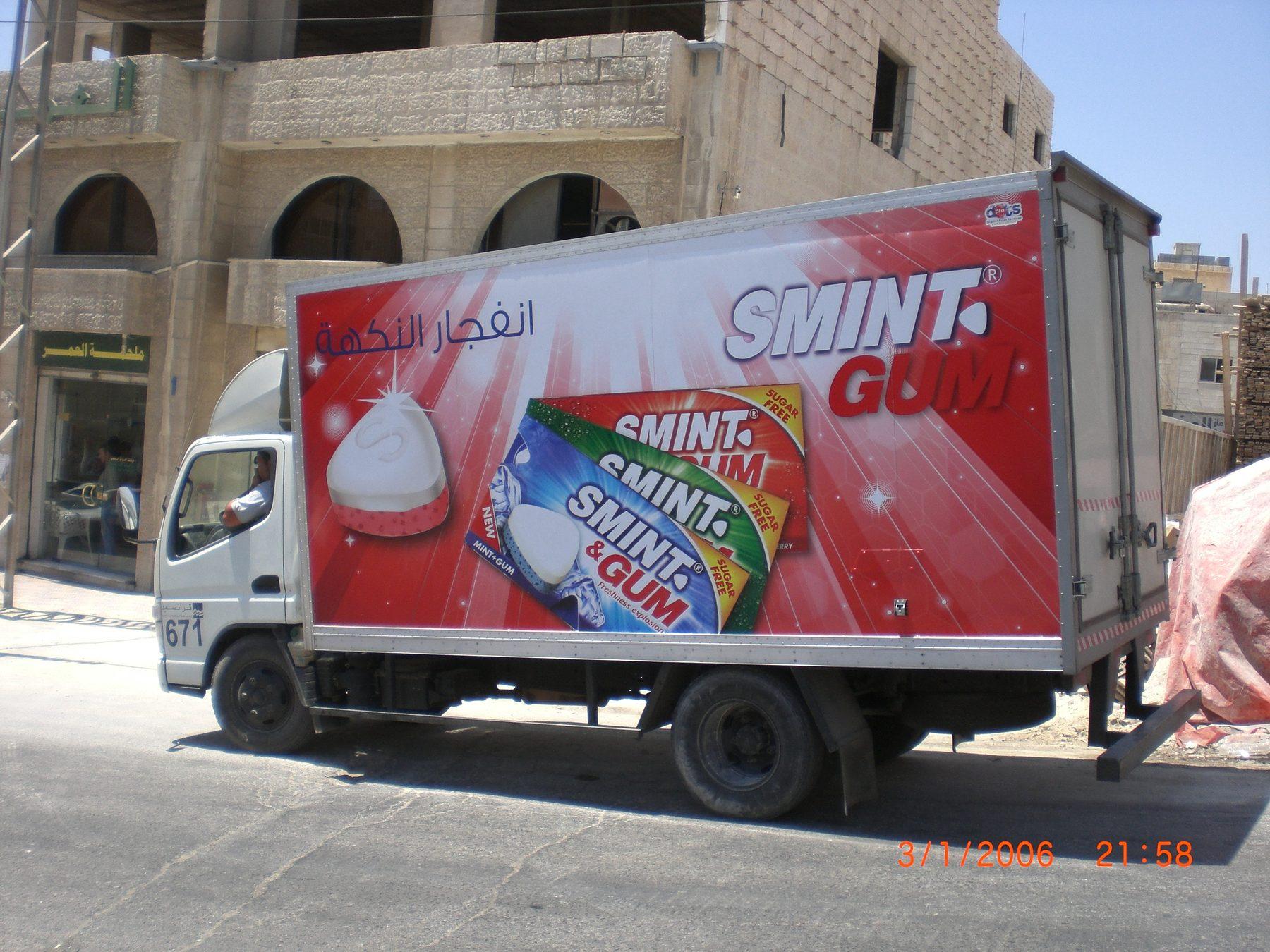 White box truck with a red wrap showing gum packs and Arabic lettering, parked on a street.