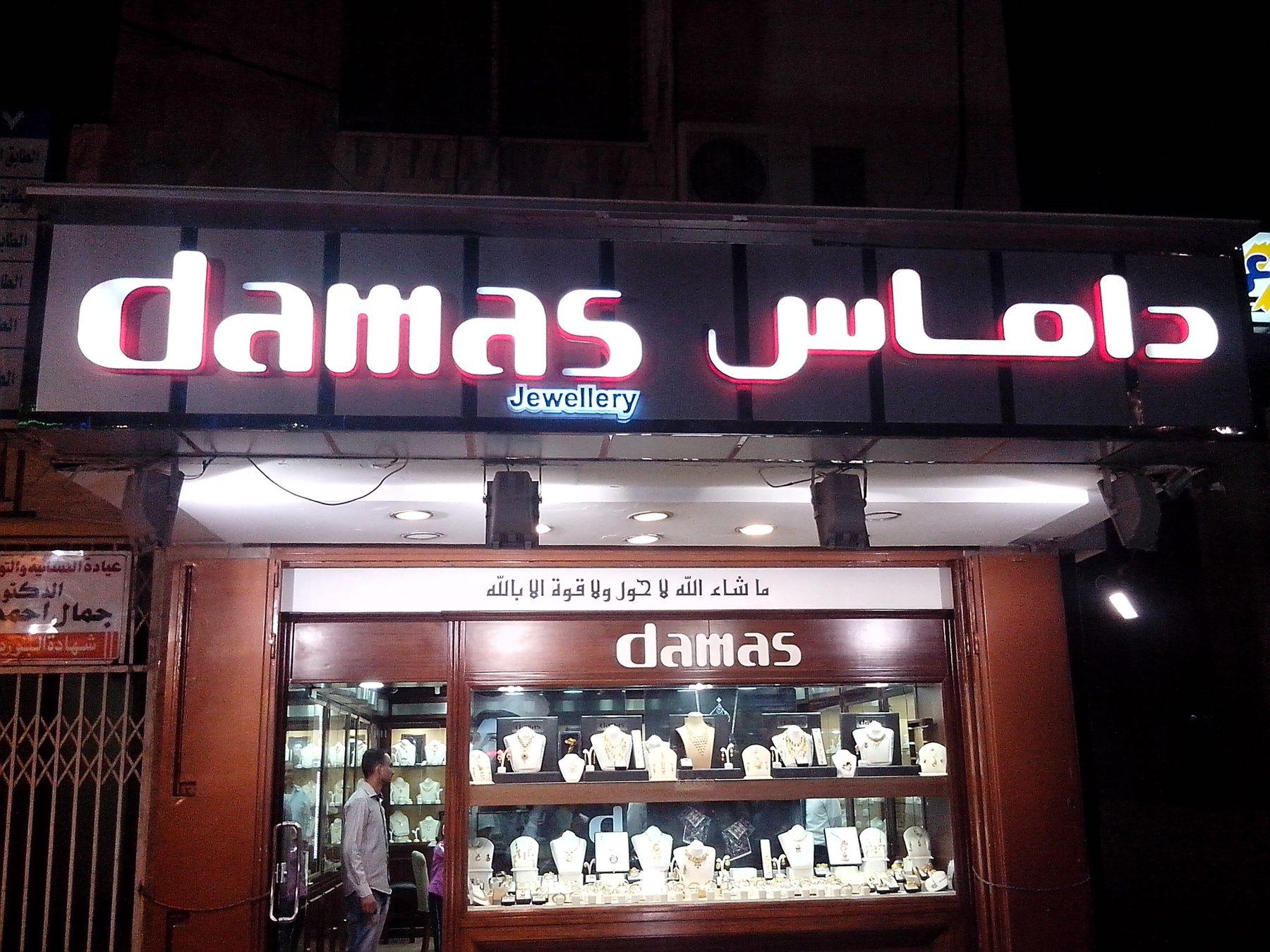 Illuminated fascia with the name in Latin and Arabic above a lit window, at dusk